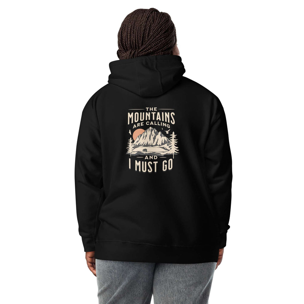 A person shown from the back wearing a black pullover hoodie with a vintage-style mountain graphic and the text 'THE MOUNTAINS ARE CALLING AND I MUST GO' printed on the back. The person is also wearing a dark knit beanie and gray sweatpants against a plain white studio background. Nature Quote Mountains Are Calling Graphic Hoodie Casual Wear Shop the Nature Quote Mountains Are Calling Graphic Hoodie, a cozy statement piece for hikers, campers, and everyday adventure seekers.